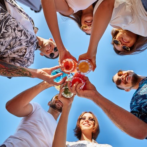 Low angle view of friends having fun at poolside summer party, clinking glasses with colorful summer cocktails near hotel swimming pool. People toast drinking fresh juice at luxury villa on vacation.