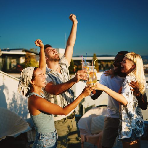Cheerful friends are raising their cocktail glasses, enjoying a rooftop party at sunset